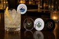 Custom logo acrylic drink tags in clear, white, and black on a bar, one clipped to a cocktail glass